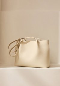 Beige Leder-Tote-Bag mit glatter Textur, doppelten langen Trägern und metallischen Akzenten; minimalistisches Design mit offenem Oberteil und ohne sichtbare Taschen.