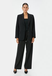 Blazer nero con chiusura a un bottone, abbinato a un crop top nero e pantaloni a palazzo. Tacchi a punta neri lucidi completano il look.