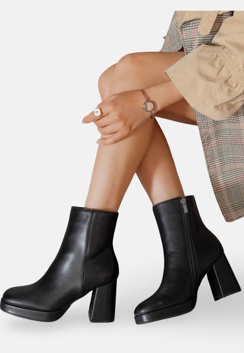 Femme portant des bottines en cuir noir à talons carrés, un manteau à carreaux beiges, une montre-bracelet et une bague, les jambes croisées sur fond blanc.