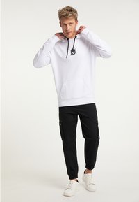 Hvid hoodie med et sort krania grafisk motiv på brystet, kengurulomme, sorte snore; parret med sorte cargobukser og hvide sneakers.