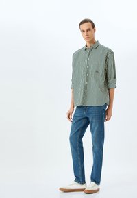 Camicia a righe verde e bianca con maniche arrotolate, abbinata a jeans blu e sneaker bianche con suola marrone. Sfondo semplice.