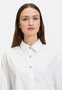 Witte blouse met een klassieke kraag, voorzien van vijf ronde, goudkleurige knopen aan de voorkant, gemaakt van een zachte stof met een subtiele glans.