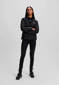 Must pufferjakk kõrge kraega, esikinnitus tõmblukuga, polsterdatud disain ja tikandiga logo. Kandmise all mustad teksad ja mustad paeltega saapad.