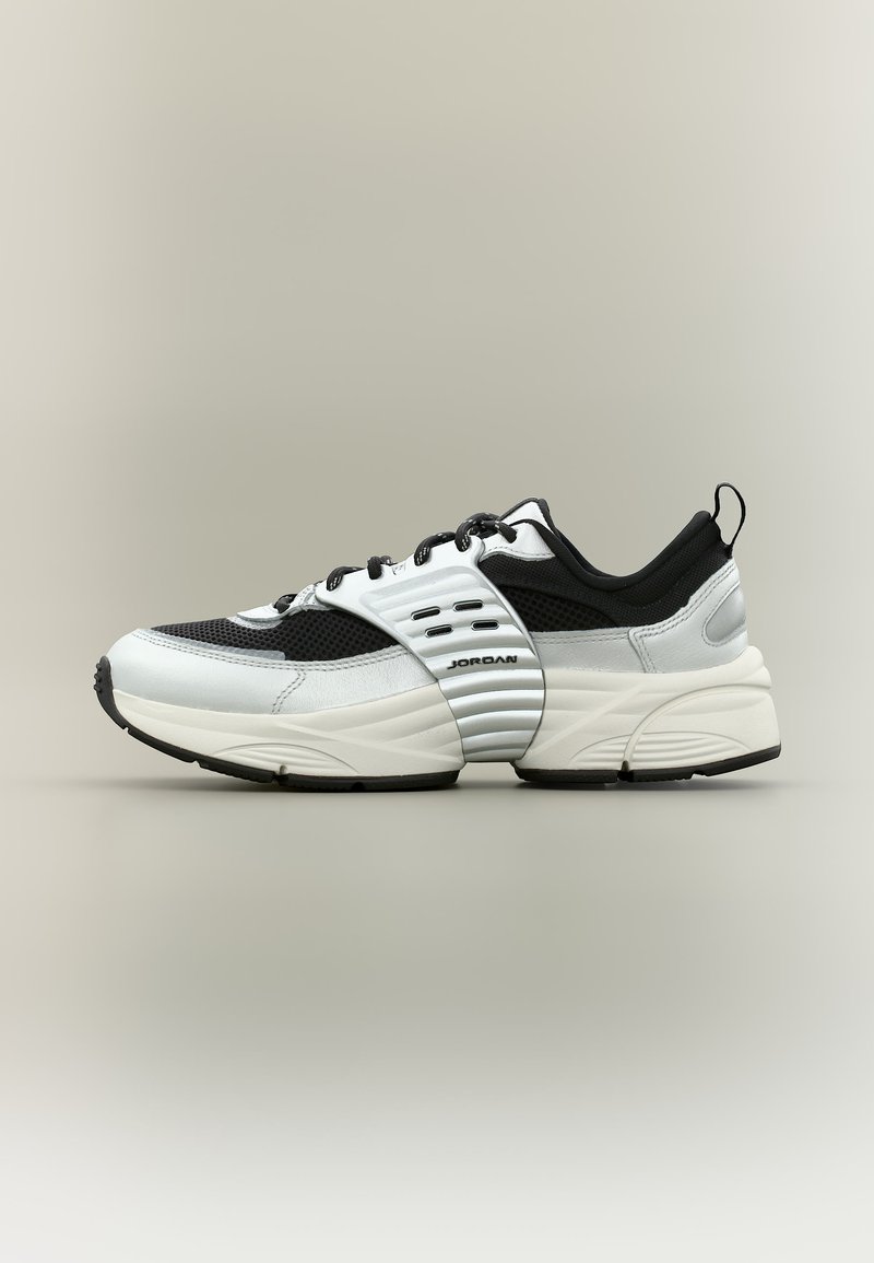 Λευκό και μαύρο χαμηλό sneaker με πάνελ από δίχτυ και δέρμα, παχιά σόλα και ριγέ λουράκι στη μέση του ποδιού με την ένδειξη "Jordan" σε ουδέτερο φόντο.