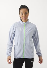 Lys grå sportsjakke med høj krave, grøn lynlås og reflekterende detaljer. Har sidelommer og fleksibel pasform for bevægelse.