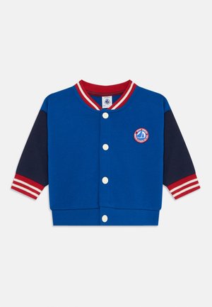 Modra trenirka s kapuco z rdečimi in belimi črtami na ovratniku in rokavih, ki ima okroglo emblem in bele gumbe.