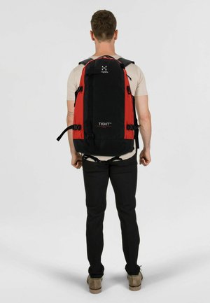 Homme portant un t-shirt beige et un pantalon noir avec un grand sac à dos noir et rouge portant l'inscription "TIGHT™", vu de dos.