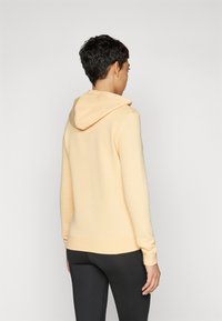 Hoodie v broskvoej farbe s jemnou textúrou, s kapucňou, dlhými rukávmi a priliehavým pásom, v kombinácii s čiernymi legínami.