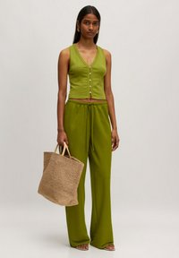 Grünes, kurz geschnittenes Tanktop mit Knöpfen, kombiniert mit weiten grünen Hosen. Das Model hält eine gewebte, beigen Tragetasche mit glatter Textur und minimalistischem Design.