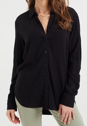 Mujer con una camisa negra holgada de botones y cuello, con varios collares dorados en capas y pantalones verde claro, de pie frente a un fondo liso.