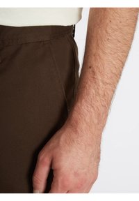 Avant-bras poilu et main d'un homme reposant à ses côtés, portant un pantalon marron foncé et une chemise de couleur crème.