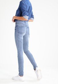 Jeans ajustados de mezclilla en azul claro con cintura alta, cinco bolsillos y cierre de cinco botones; combinados con una camisa azul de botones.