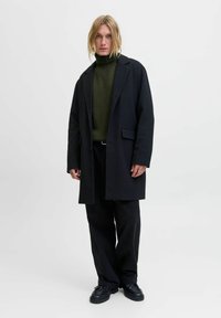 Schwarzer oversized Mantel mit aufgeschlagenem Kragen, strukturiertem olivgrünem Rollkragenpullover und lockeren schwarzen Hosen, kombiniert mit schwarzen Schuhen.