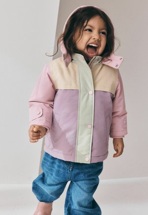 Junges Kind trägt eine pink-beige Jacke und blaue Jeans, lächelt breit mit offenem Mund und steht vor einem schlichten Hintergrund.