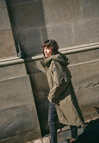 Vrouw in een olijfgroene parka met capuchon en zwarte broek die naast een getextureerde stenen muur in het zonlicht loopt.