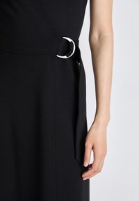 Zbliżenie prawego ramienia osoby obok czarnej sukienki z materiałowym paskiem przymocowanym srebrną klamrą D-ring w talii.