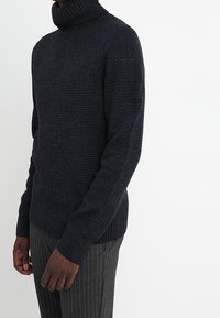 Navyblå stickad polotröja med en strukturerad mönstring, åtsittande ärmar och ribbad nederkant, bäddad över randiga byxor.