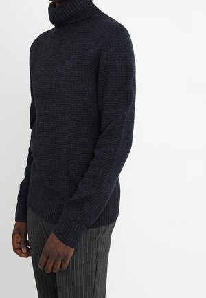 Dunkelblauer gestrickter Rollkragenpullover mit strukturiertem Muster, taillierten Ärmeln und geripptem Saum, getragen über gestreiften Hosen.