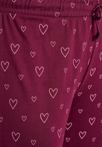 Burgundinpunainen kangas, jossa on pehmeä tekstuuri ja vaaleanpunaisia sydämiä hajanaisesti kuvioituna. Venyvä vyötärönauha ja säädettävä nyöri.