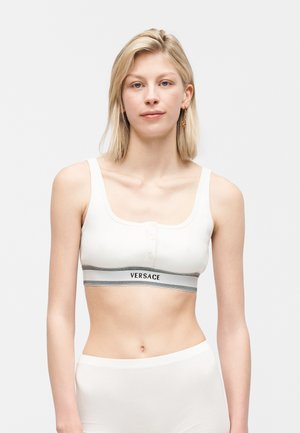 Blondynka w białym, przyciskanym topie Versace, stojąca na tle jednolitego, jasnego tła, zwrócona przodem, z neutralnym wyrazem twarzy.