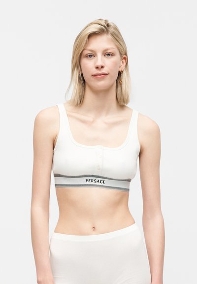 Ξανθιά γυναίκα που φοράει λευκό crop top Versace με κουμπιά, στέκεται μπροστά σε απλό φωτεινό φόντο, κοιτάζει μπροστά με ουδέτερη έκφραση.