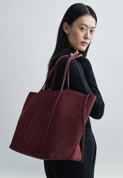 Burgundy suede tote bag with long, thin straps; features a structured shape and minimalistic design. No visible logos or embellishments.
