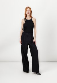 Black sleeveless top paired with high-waisted, wide-leg pinstriped trousers. The model holds a small black handbag and wears black pointed heels.