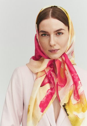 Femme à la peau claire et aux cheveux bruns porte un blazer rose et un foulard coloré noué autour de sa tête avec des motifs rouges, jaunes et crème.