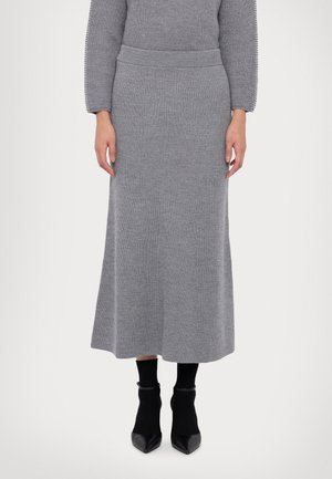 Kobieta ubrana w długą, szarą, prążkowaną dzianinową spódnicę, pasujący szary prążkowany sweter, czarne skarpetki do kostek i czarne szpiczaste szpilki na białym tle.