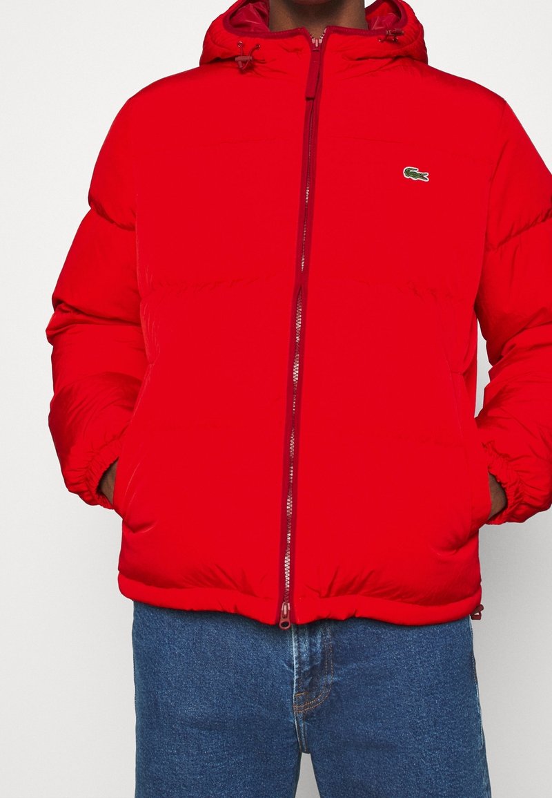 Veste matelassée rouge avec fermeture éclair avant, capuche, poignets élastiques et deux poches latérales. Présente un petit logo brodé sur la poitrine.