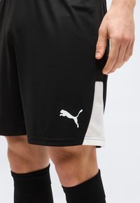 Μαύρα αθλητικά σορτς με λευκές πλευρικές επενδύσεις από δίχτυ και λευκό λογότυπο Puma, φορεμένα με μαύρες κάλτσες μέχρι το γόνατο και με ορατό χέρι που ακουμπάει στο μηρό.