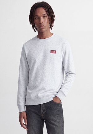 Jeune homme avec des dreadlocks portant un sweat-shirt gris clair avec un patch logo rouge Jack & Jones et un jean noir, debout avec une main dans la poche.