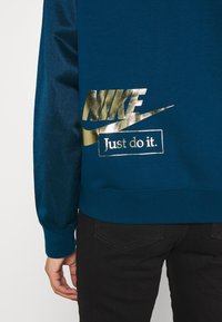 Personne portant un sweat-shirt bleu avec un logo Nike doré brillant et le texte "Just do it." en bas du dos, associé à un pantalon noir.