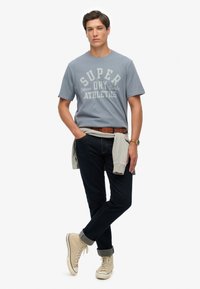 Camiseta de color azul claro con gráfico "SUPER", jeans oscuros, zapatillas beige y una sudadera gris atada a la cintura. Conjunto informal.