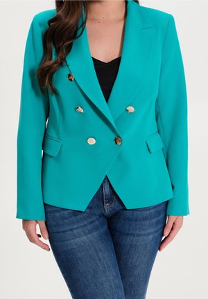 Femme portant un blazer bleu sarcelle croisé avec des boutons dorés, sur un haut noir et un jean bleu, debout devant un fond uni.