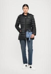 Vrouw in een zwarte gewatteerde jas, spijkerbroek, witte sneakers, met een blauwe handtas tegen een effen lichte achtergrond.