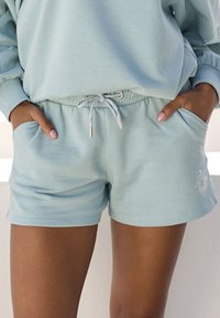 Gaiši zili kokvilnas šorti ar elastīgu jostu, auklu ar zelta akcentiem, sānu kabatām un dekoratīvu logo uz apakšmalas.