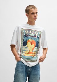 Balta per didelė t-shirt su spalvinga grafika, vaizduojančia sėdinčią kobrą, apsupta dekoratyvinio rašto, derinama su mėlynomis džinsinėmis kelnėmis.