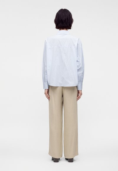 Personne aux cheveux courts et foncés, de dos, portant une chemise bleu clair à manches longues et un pantalon beige à jambes larges avec des chaussures foncées.