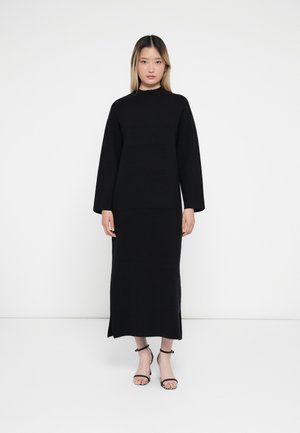 Black long-sleeve dress with a high neckline, made of a smooth fabric. Features a straight cut and side slit. Worn with strappy black sandals.