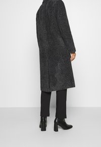 Cappotto grigio scuro, testurizzato, con taglio dritto, maniche lunghe e spacchi laterali, abbinato a stivali neri con tacco alto e pantaloni slim.