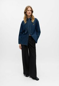 Camicia di jeans blu scuro con colletto annodato, pantaloni neri larghi e calzature nere. Presenta un logo discreto sulla camicia e una vestibilità comoda.
