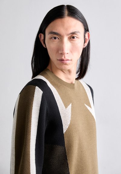 Olíva zöld testtel, fekete és fehér geometrikus mintákkal, valamint texturált díszítésekkel rendelkező kötött pulóver.