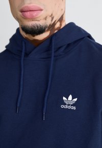Marinblå huva med en vit Adidas-logotyp på bröstet. Tyget ser mjukt ut, med synliga dragsnören och en känguruficka.