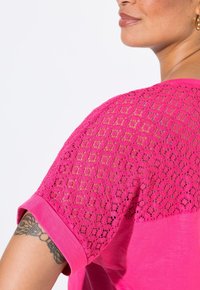 Růžová košile s pevným předním dílem a krajkovým zadním panelem. Krátké rukávy s ohrnutými manžetami, které vykazují texturovaný design.