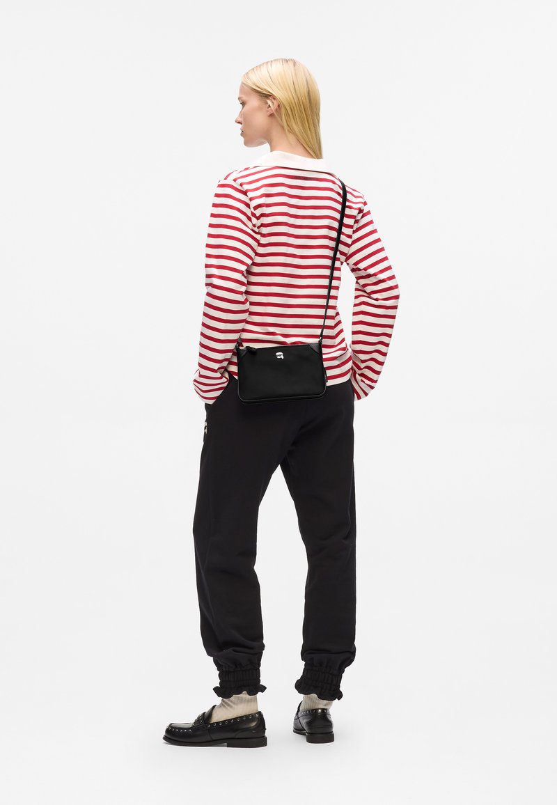 Borsa a tracolla nera con una texture liscia e un design minimalista, abbinata a una maglietta a maniche lunghe rossa e bianca a righe e pantaloni jogger neri.