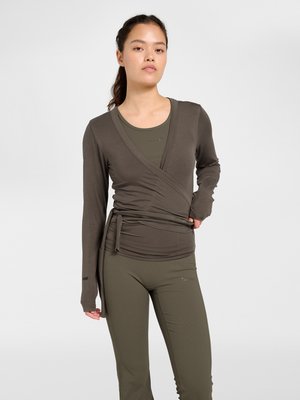 Frau trägt ein olivgrünes Wickel-Top mit langen Ärmeln und passende enganliegende Leggings, steht vor einem schlichten hellen Hintergrund.