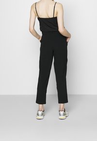 Svart ärmlös topp med tunna axelband, kombinerad med svarta snäva byxor. Modellen bär grå och gula sneakers. Enkel, minimalistisk design.