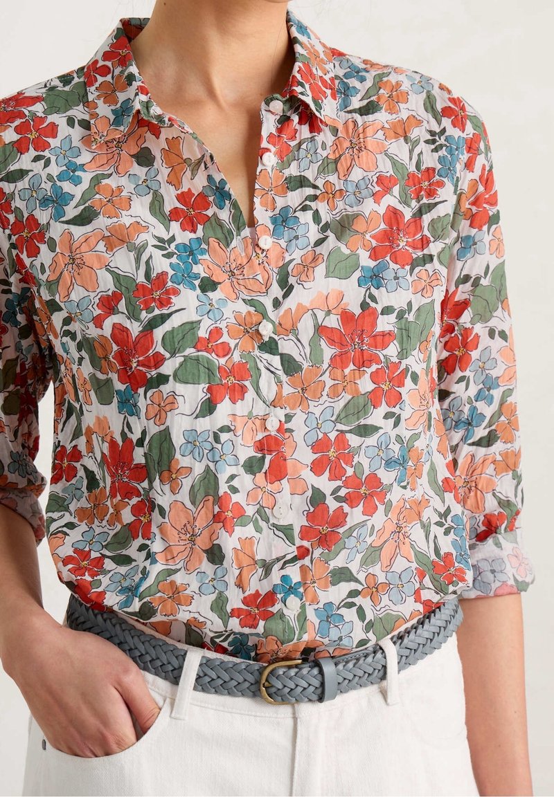 Femme portant une chemise à fleurs boutonnée avec des fleurs orange, rouges, bleues et des feuilles vertes, rentrée dans un pantalon blanc avec une ceinture tressée grise.