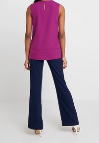 Fioletowy bezrękawnik z dekoltem w kształcie łezki, w połączeniu z szerokimi granatowymi spodniami. Top ma gładką fakturę; spodnie są lekko marszczone.